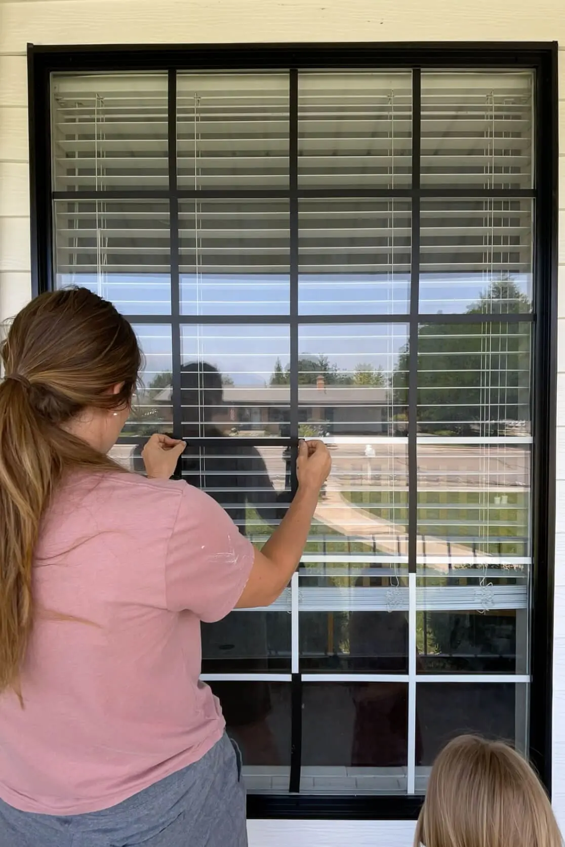 Woman pressing black vinyl strips onto horizontal grid lines of a window to create a modern black grid design.